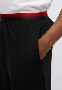 Pantalones deportivos negros con una textura suave, que cuentan con una cinturilla elástica ancha que pasa de rojo a negro. Incluyen bolsillos laterales.