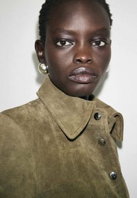 Veste en daim vert olive avec un col, ornée de grands boutons en métal et d'une texture douce, présentée sur un fond neutre.