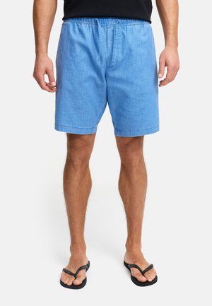 Mężczyzna ubrany w jasnoniebieskie spodenki z sznurkiem i zamkiem błyskawicznym oraz czarne klapki, stojący na jednolitym tle.