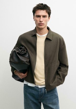 Junger Mann in olivgrüner Jacke, cremefarbenem Pullover und blauen Jeans hält eine große schwarze Ledertasche unter dem Arm vor einem schlichten Hintergrund.