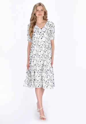 Ung kvinde med langt blondt hår iført en hvid blomsterprintet knælængde kjole og hvide sandaler med hæl, stående mod en hvid baggrund.