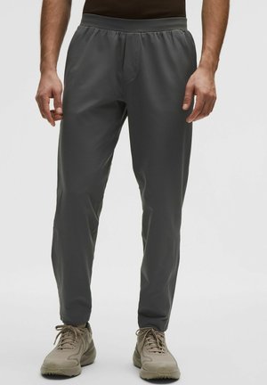 Mand iført mørkegrå elastiske bukser med talje og beige træningssko, stående mod en ensfarvet hvid baggrund.