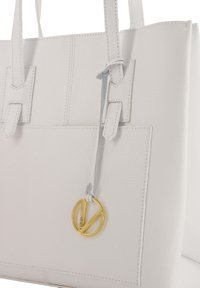 Biała skórzana torba tote z teksturowaną powierzchnią, podwójnymi uchwytami i złotą zawieszką z logo na szarym pasku. Minimalistyczny design bez dodatkowych akcentów.
