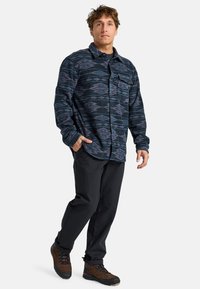 Camicia di flanella blu scuro con motivi geometrici in viola e rosso, abbinata a pantaloni neri e scarponi da trekking marroni.