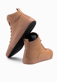 Hnědé vysoké tenisky s hladkým syntetickým materiálem, zaoblenou špičkou a tónovými tkaničkami. Mají texturovanou gumovou podrážku a embosované logo.