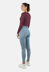 Bordeauxrotes, langärmeliges, kurzes Oberteil kombiniert mit hellblauen, hoch geschnittenen Jeans. Das Model trägt weiße Sneakers und zeigt eine figurbetonte Silhouette.