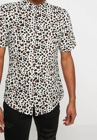 Camicia a maniche corte in tessuto bianco con stampa leopardata nera e marrone, dotata di colletto a punta e chiusura con bottoni frontali.