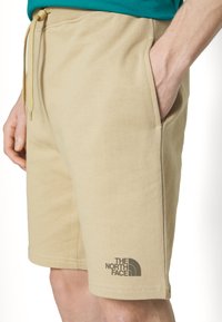 Pantaloni corti beige in cotone con tasche laterali e chiusura con cordoncino. Presenta il logo The North Face stampato in grigio scuro sull'orlo sinistro.