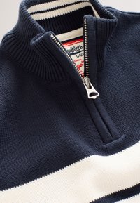 Námornícky modrý pletený pulóver s vysokým golierom a štvrťovým zipsom. Má biely horizontálny pruh a rebrovanú textúru pozdĺž goliera a spodného lemu.