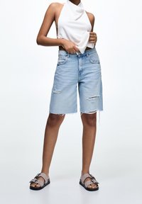 Pantaloni shorts in denim blu chiaro con orlo sfrangiato e dettagli strappati. Abbinati a un top bianco a halter e sandali grigi con fibbia.