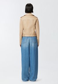 Chaqueta de moto de cuero beige con una silueta estructurada, cuello alto y cinturón en la cintura, combinada con vaqueros de denim de pierna ancha en azul claro.