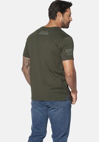 Olive green short-sleeve t-shirt with a crew neck, featuring handwritten text and logos on the back and sleeve. Cotton material, regular fit.