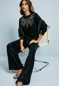 Felpa nera con borchie dorate decorative e maniche frangiate, abbinata a pantaloni neri a palazzo in paillettes. Modella seduta su una sedia in cromo.