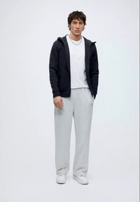 Jonge man staand, met witte sneakers, lichtgrijze broek, wit T-shirt en zwarte zip-up hoodie, tegen een effen witte achtergrond.