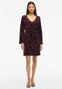 Vīna sarkanā spīdīgā kleita ar garām piedurknēm un V veida izgriezumu. Īss garums, pieguļošs siluets, ar teksturētu spīdīgu rakstu visā kleitā.