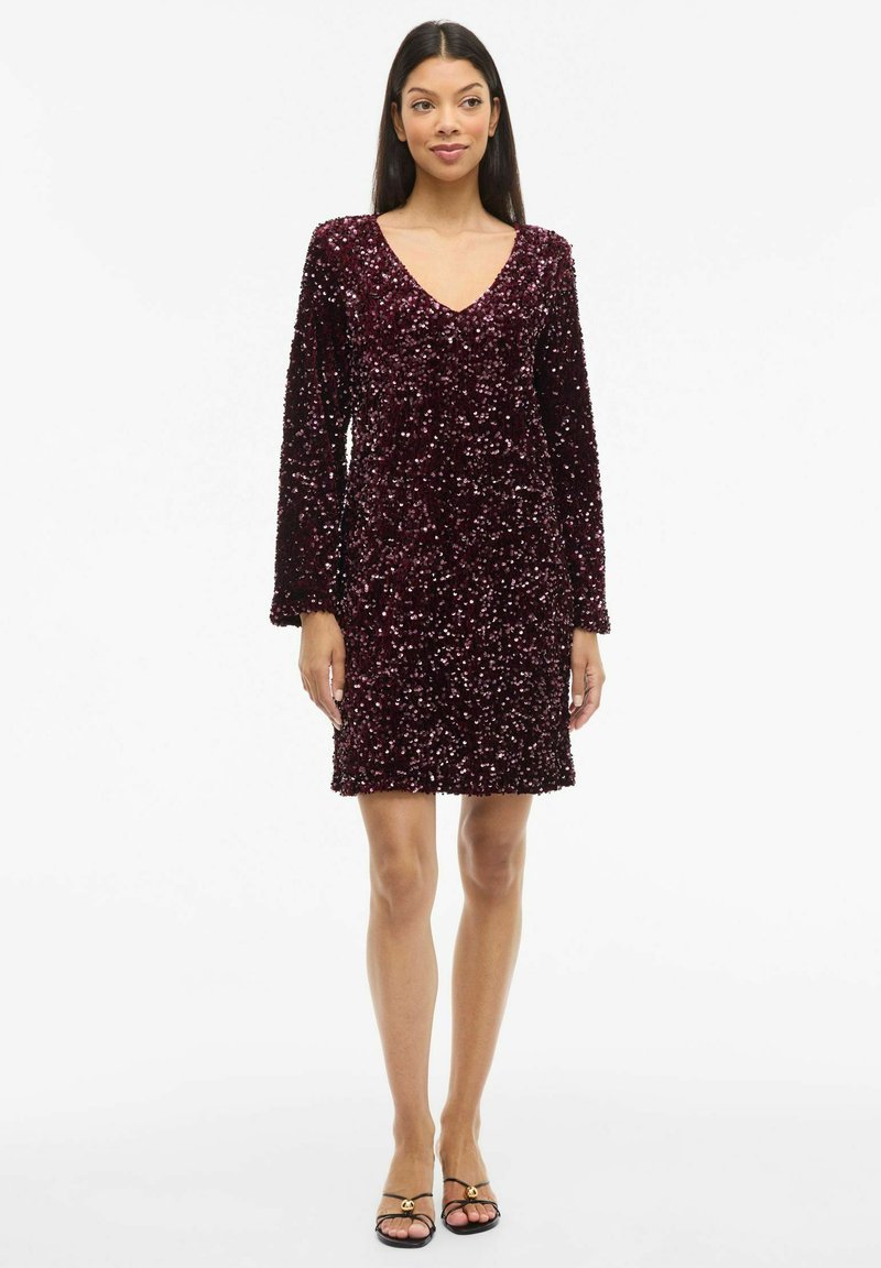 Vīna sarkanā spīdīgā kleita ar garām piedurknēm un V veida izgriezumu. Īss garums, pieguļošs siluets, ar teksturētu spīdīgu rakstu visā kleitā.