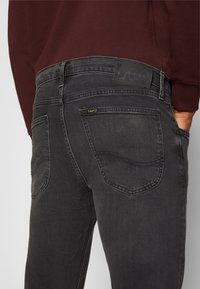Nahaufnahme von dunkelgrauen Slim-Fit-Jeans mit Gesäßtaschen und schwarzem Lee-Markenetikett, getragen mit einem marsala-farbenen Langarmshirt.