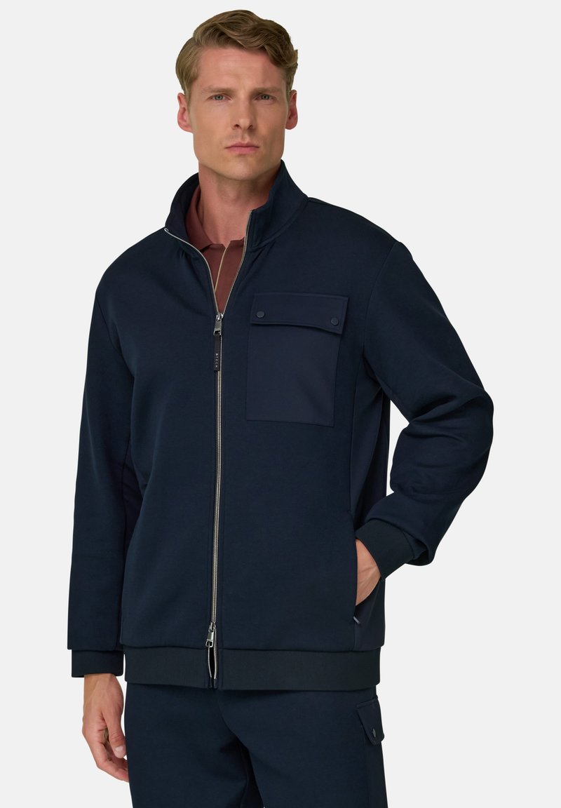 Homme portant une veste zippée bleu marine foncé avec une poche poitrine boutonnée et un pantalon assorti, debout avec une main dans la poche.
