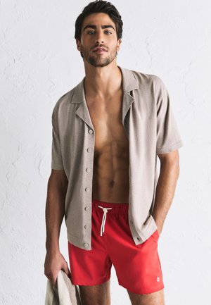 Jonge man die een open beige korte mouwen overhemd en rode zwembroek draagt, houdt een handdoek tegen een witte gestructureerde muur.