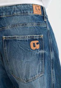 Jeans in denim blu con una finitura texture, caratterizzati da un'etichetta in pelle con logo, cuciture a vista e un design della tasca posteriore.