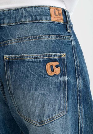 Jean en denim bleu avec une finition texturée, comportant une patch en cuir avec logo, des coutures visibles et un design de poche arrière.