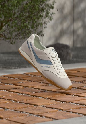 Valkoinen ja sininen Valentino-tennari kumipohjalla, kelluu teksturoidun puisen ja kivisen ulkopinnan yläpuolella, taustalla sumeaa vihreyttä.