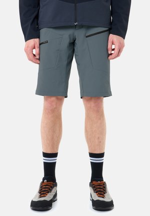 Uomo che indossa pantaloncini grigi con cerniera, calzini neri con strisce bianche e scarpe da trekking grigie con lacci arancioni, in piedi contro uno sfondo bianco.