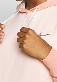 Händer justerar de vita dragbanden på en ljusrosa Nike-hoodie, vilket visar en del av halslinjen och logotypen på bröstet.