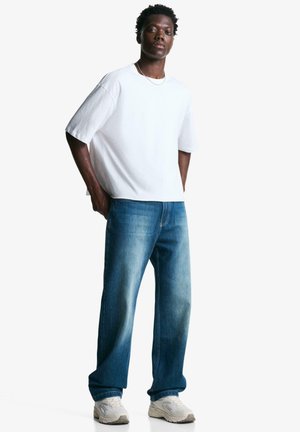 Witte katoenen t-shirt met korte mouwen, gecombineerd met blauw vervaagde straight-leg jeans. Model draagt witte sneakers en staat tegen een effen achtergrond.