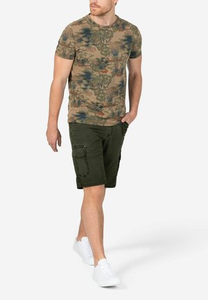 T-Shirt mit Camouflage-Muster in Beige, Grün und Blau. Kombiniert mit olivgrünen Cargo-Shorts, getragen mit weißen Sneakers.