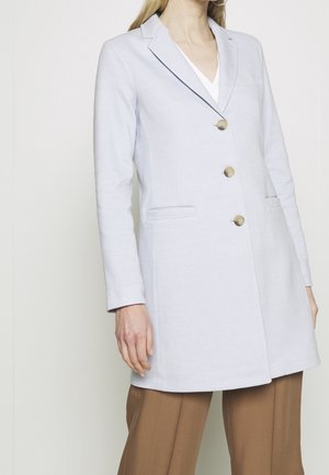 Femme portant un long blazer gris clair avec trois boutons et un pantalon marron sur fond blanc uni.