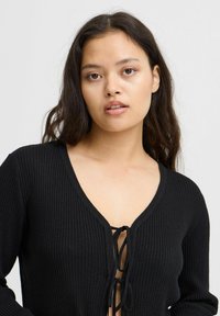 Svart ribbad cardigan med djup V-ringning och knytning framtill. Långa ärmar och mjukt material förbättrar dess bekväma design.