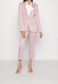 Suit in un delicato rosa a scacchi, composto da una giacca doppiopetto e pantaloni a gamba larga accorciati, abbinato a una camicia bianca con fiocco e tacchi trasparenti.