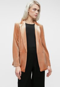 Femme aux longs cheveux blonds portant un blazer en velours beige sur un haut noir et un pantalon noir, se tenant contre un fond clair.