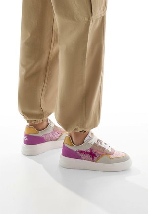Chunky sneakers feature a white sole, pink and purple glitter accents, gray and yellow panels, and white laces. Worn with beige joggers.