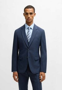 Navy blue suit with a subtle check pattern, single-breasted jacket, notch lapels, and two buttons. Paired with a light blue shirt and striped tie.