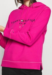 Růžová mikina s kapucí, stahovacími šňůrkami, nápisem s logem a barevnými bloky. Má volný střih a šití na lemu a manžetách.