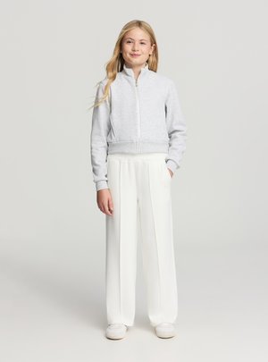 Felpa grigia con zip e orlo a coste, abbinata a pantaloni bianchi a gamba larga. Modella in piedi con le mani nelle tasche, indossa sneakers chiare.