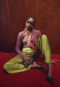 Blusa de encaje rosa suave con detalles de volantes, combinada con pantalones brillantes verdes oliva. Unos tacones negros de tiras y un clutch negro completan el atuendo.