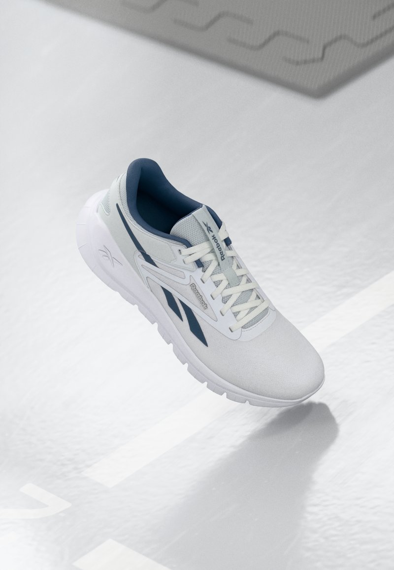 White athletic shoe with dark blue accents, textured fabric upper, and thick rubber sole. Features lace-up design and logo detail on side.