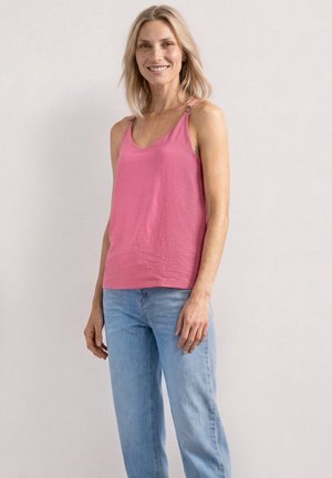 Femme souriante aux cheveux blonds portant un haut rose sans manches avec des fermoirs métalliques et un jean bleu clair, debout devant un fond uni.