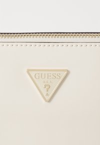 Cremefarvet pung med et trekantet guldlogo med "GUESS U.S.A." og et spørgsmålstegn, afsluttet med en guldlukning.