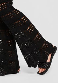 Mustad kudumislaiusega laiad püksid, millel on aukudega muster, koos mustade põimitud rihmadega varbavahetega sandaalidega heledal taustal.