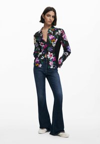 Camicia a stampa floreale con base nera, maniche lunghe e colletto a punta. Abbinata a jeans a zampa d'elefante blu scuro e scarpe chiare.