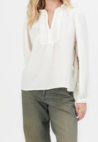 Biała bluzka z długim rękawem, z falbaniastym dekoltem i dekoracyjnymi zakładkami, w połączeniu z jasnorozowym denimowymi spodniami. Miękka, teksturowana tkanina.