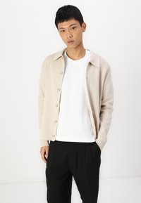 Beige button-up cardigan with a relaxed fit, round collar, and white T-shirt underneath, paired with black pleated trousers.