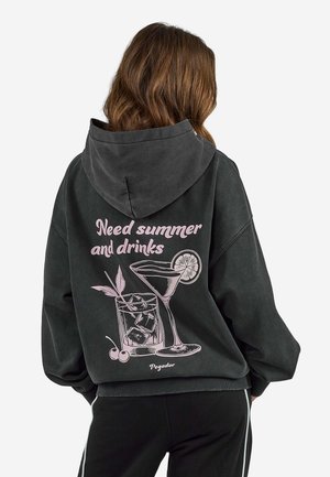 Osoba o brązowych włosach nosząca ciemną bluzę z kapturem z różową ilustracją napojów i tekstem "Need summer and drinks" na plecach.