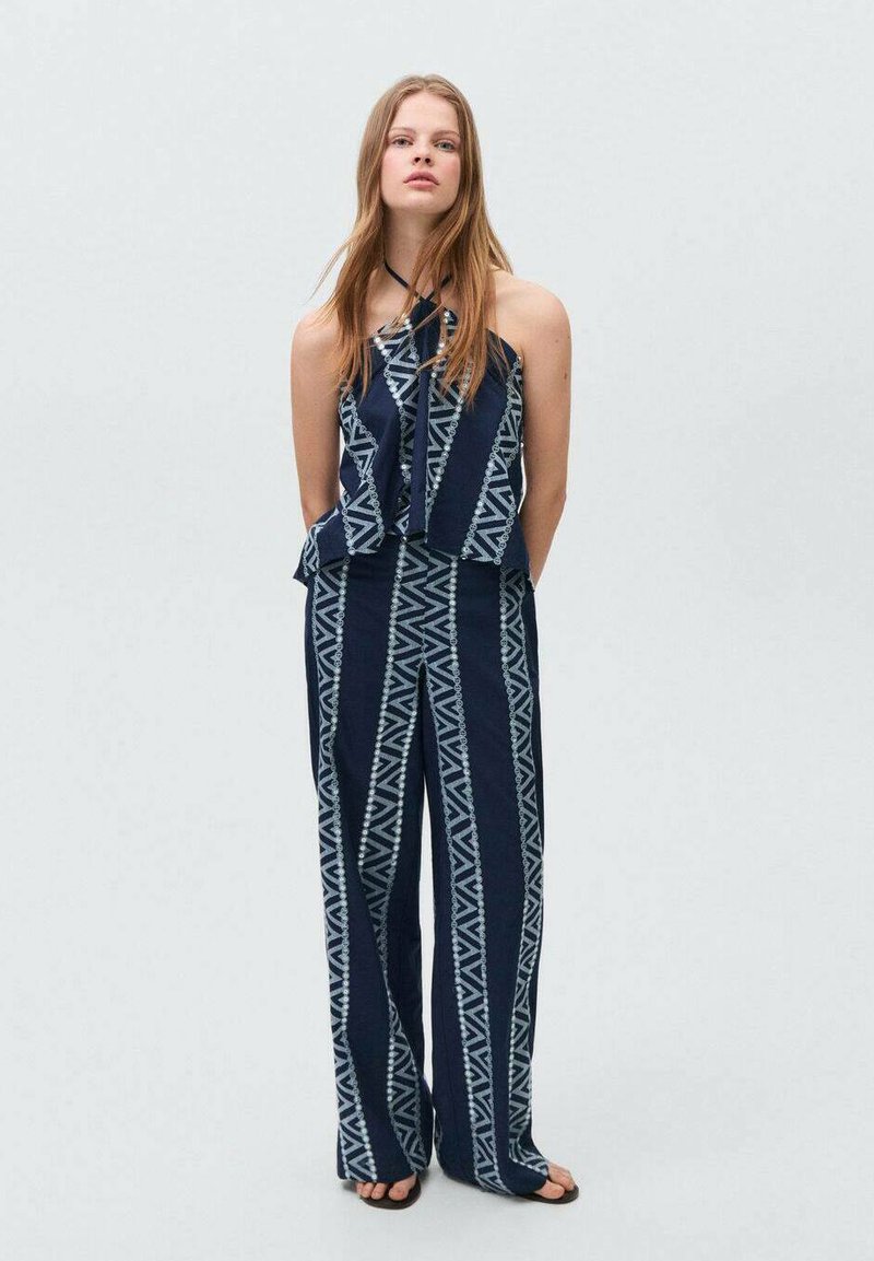 Marineblå og hvidmønstret jumpsuit med halterneck, brede ben og geometriske designs. Stoffet ser glat ud med en let tekstur.