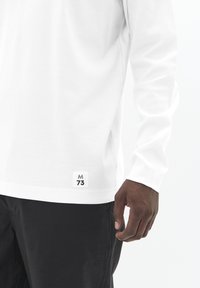 Vyras vilkintis baltą ilgam rankovėms marškinį su maža etikete "M 73" šalia krašto, derinamas su juodomis kelnėmis, priešais vientisą foną.