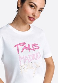 Tricou alb cu mânecă scurtă, decorat cu strassuri colorate, având textul "PARIS MADRID ITALY" în nuanțe de roz, aurie și neagră.